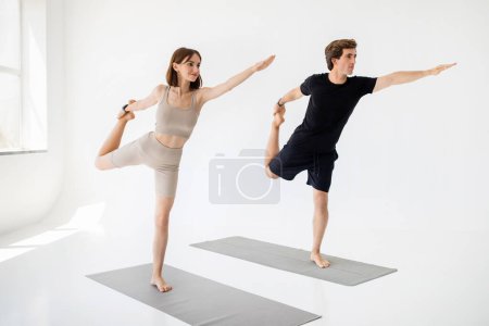 Téléchargez les photos : Souriant jeune homme et femme européens en vêtements de sport faire des exercices d'étirement, pratiquer le yoga, profiter d'un mode de vie actif sur tapis dans le salon ou salle de gym intérieure. Soins du corps, entraînement ensemble et sport - en image libre de droit