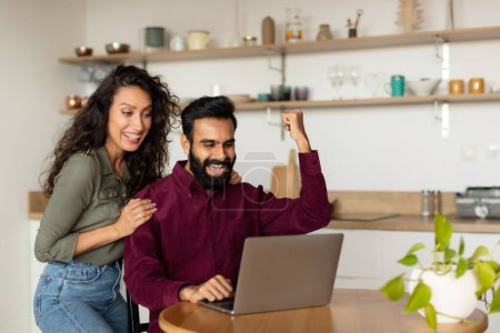 Téléchargez les photos : Émotionnel joyeux conjoints arabes serrant les poings et criant devant un ordinateur portable dans la cuisine, heureux homme et femme du Moyen-Orient regardant l'écran et le jeu ensemble, copier l'espace - en image libre de droit