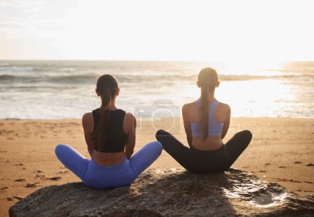 Téléchargez les photos : Les jeunes femmes européennes jumelles en vêtements de sport pratiquent le yoga et des exercices de respiration en position lotus, profitent du lever du soleil sur la plage de la mer, en plein air, dos. Entraînement ensemble, mode de vie actif et soins corporels matinaux - en image libre de droit