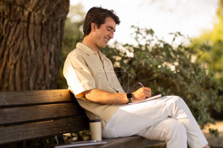Téléchargez les photos : Beau jeune homme créatif portant un écrivain décontracté en tenue d'été assis sur un banc dans un parc public, prenant des notes sur un bloc-notes et souriant. Blogueur travaillant à l'extérieur, écrivant des idées, copiant de l'espace - en image libre de droit