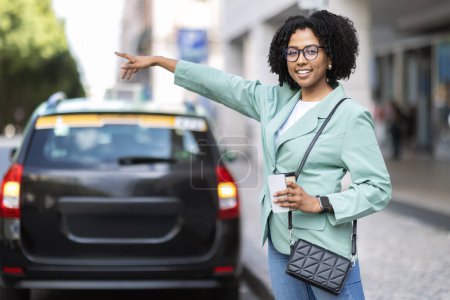 Photo for Attractive young black woman wearing smart casual outfit with takeaway coffee and smartphone in her hand standing on street, going to office, catching taxi, raising hand up, copy space - Royalty Free Image