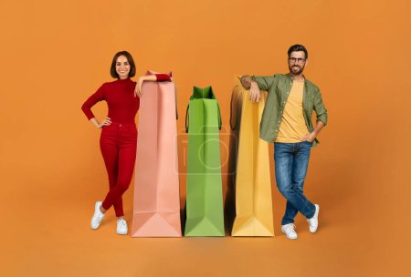 Photo for Shopping, Sale Season. Happy Buyers Couple Leaning On Big Paper Shopper Bags, Advertising Discounts And Smiling At Camera Standing Over Orange Background In Studio. Collage, Full Length Shot - Royalty Free Image