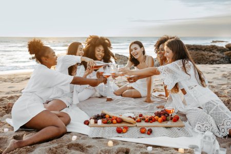 Foto de Fiesta de despedida de soltera. Feliz diversas mujeres abriendo botella de champán, teniendo picnic en la playa de arena en la costa, espacio libre. Noche de soltera, soltera - Imagen libre de derechos