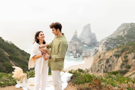 Téléchargez les photos : Beau couple européen amoureux ouvrant une bouteille de champagne, ayant rendez-vous romantique sur une falaise rocheuse avec belle vue sur la côte océanique, espace libre - en image libre de droit
