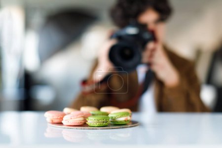 Photo for Man food influencer photographer taking photos of sweet tasty macarons in photo studio, holding digital camera, selective focus on confectionery - Royalty Free Image