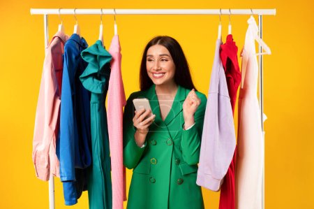 Photo for Overjoyed shopaholic lady using cellphone and shaking fist, standing near clothing rail and reading news about sales and discounts, yellow studio background - Royalty Free Image