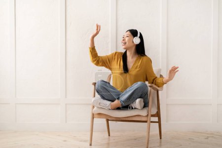 Foto de Alivio del estrés, relajación en casa. Alegre joven asiática feliz sentada en sillón, usando auriculares inalámbricos, escuchando música con los ojos cerrados, cantando y bailando, espacio para copiar - Imagen libre de derechos