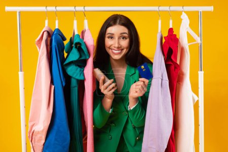 Photo for Cheerful young shopaholic lady buying clothes online, woman customer holding cellphone and credit card, posing near clothing rail over yellow studio background - Royalty Free Image