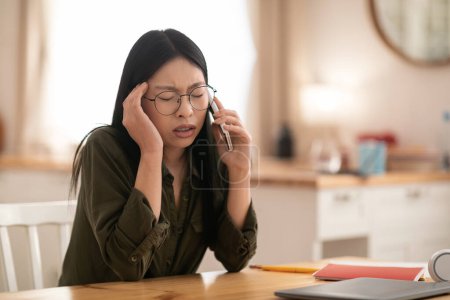 Photo for Distressed beautiful millennial asian businesswoman in comfy casual outwear and eyeglasses sitting at kitchen, having phone conversation, touching head, copy space for ad, have troubles in business - Royalty Free Image