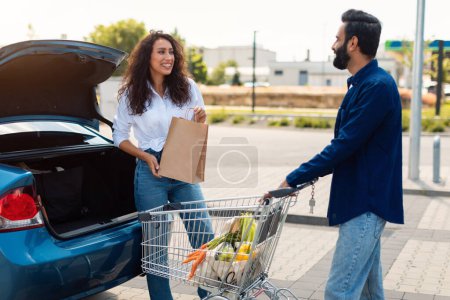 Téléchargez les photos : Heureux couple arabe emballage sacs d'épicerie dans un coffre de voiture sur un parking, femme regardant et parlant avec son petit ami, homme debout près du panier plein de nourriture fraîche - en image libre de droit