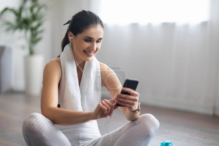 Photo for Music for fitness, mobile app. Smiling beautiful sporty young woman sitting on yoga mat, using smartphone and earphones, listening to music while exercising at home, copy space - Royalty Free Image