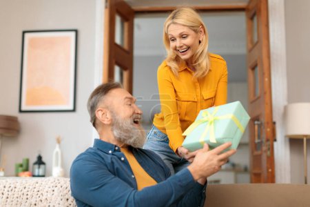 Loving mature woman surprising her husband with wrapped gift at home. Blonde wife greeting partner on birthday or anniversary, giving present box, celebrating valentines day and family holiday