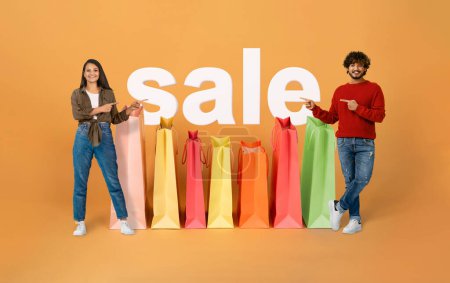 Photo for Shopping Offer. Happy Indian Buyers Couple Pointing At Word Sale And Big Paper Shopper Bags Standing On Orange Studio Background, Collage. Spouses Shopaholics Advertising Discounts. Full Length - Royalty Free Image