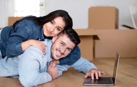 Photo for Cheerful Couple Relaxing With Laptop After Moving Home Lying On Floor, Smiling Young Spouses Using Computer Together, Purchasing Furniture Online Posing Among Carton Boxes Indoor - Royalty Free Image