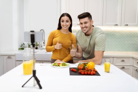 Photo for Happily married happy young couple famous food bloggers cooking healthy dinner fresh vegetable salad and streaming, using smartphone set on tripod, showing thumb ups, modern kitchen interior - Royalty Free Image