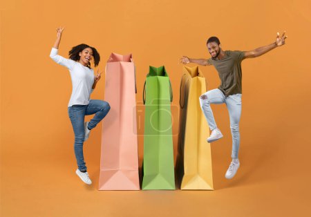 Photo for Consumerism. Joyful Shopaholics Black Couple Jumping Posing Near Large Paper Shopping Bags, Advertising Sale Offer On Orange Studio Background, Smiling At Camera. Discount Advertisement. Collage - Royalty Free Image