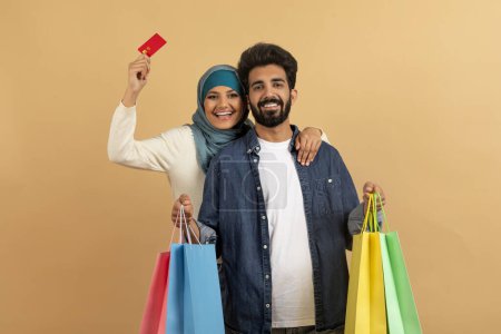 Téléchargez les photos : Easy Shopping. Heureux couple islamique avec carte de crédit et sacs à provisions en papier souriant à la caméra tout en posant ensemble sur fond de studio beige, conjoints arabes appréciant faire des achats, espace de copie - en image libre de droit