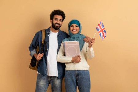 Téléchargez les photos : Étudier à l'étranger. joyeux jeunes étudiants arabes couple avec sacs à dos tenant drapeau britannique, heureux sourire musulman et femme dans le hijab montrant Union Jack tout en posant sur fond de studio beige - en image libre de droit