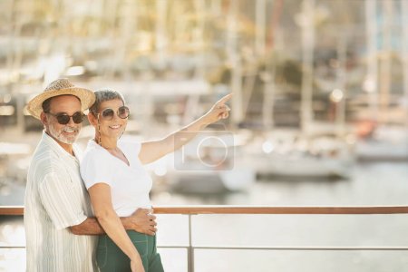 Photo for Look There. Joyful Mature Tourists Couple Embracing And Pointing Finger Aside At Backdrop Of Luxury Sailboats At Sea Marina, Standing Outdoor And Smiling At Camera. Empty Space For Text - Royalty Free Image