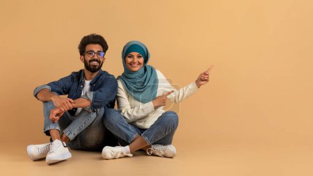 Foto de Mira esto. Jóvenes cónyuges árabes señalando a un lado en el espacio de copia, el hombre musulmán y la mujer en Hijab mostrando lugar libre para su anuncio u oferta, sentados juntos en un fondo beige, Panorama - Imagen libre de derechos
