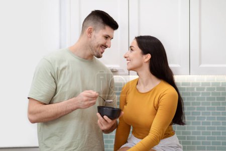 Photo for Beautiful young couple in love have breakfast together at home. Happy handsome millennial man feeding his pretty long-haired girlfriend or wife with oatmeal, kitchen interior, copy space - Royalty Free Image