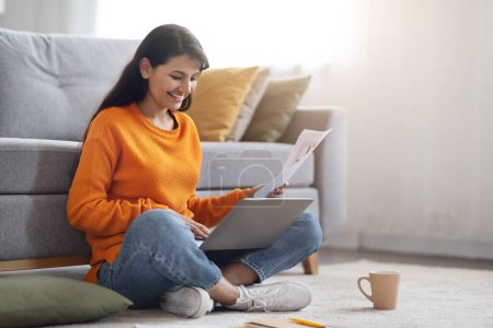 Photo for Smiling beautiful young indian woman wearing casual outfit freelancer working from home, sitting on floor by couch, using laptop computer, holding papers, typing on pc keyboard, copy space - Royalty Free Image