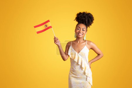 Foto de Alegre joven afroamericana mujer en vestido sostienen bandera de España, aislado en el fondo del estudio amarillo. Intercambio de estudios, aprendizaje de idiomas, estilo de vida, educación, publicidad y oferta - Imagen libre de derechos