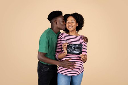 Photo for Happy young african american husband kissing cheek pregnant wife with big belly in casual, show ultrasound shot, isolated on beige studio background. Medical exam, waiting for baby, love, family - Royalty Free Image