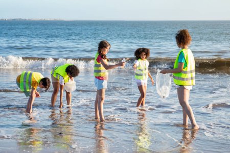 Téléchargez les photos : Les adolescents sérieux les élèves multiethniques bénévoles dans les gilets recueillent les ordures et les bouteilles dans l'océan, travaillent sur la plage, toute la longueur. Eco care, sauvegarde de l'environnement, recyclage du plastique et travail d'équipe - en image libre de droit