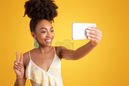 Photo for Cheerful young african american lady in dress taking selfie on smartphone, make peace sign, isolated on yellow studio background. Lifestyle, fashion blog, ad and offer, gadget for photo - Royalty Free Image