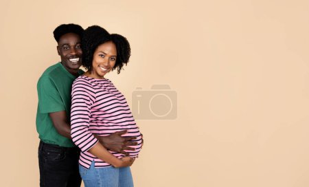 Téléchargez les photos : Souriant jeune mari afro-américain étreignant femme enceinte dans un cadre décontracté avec gros ventre, isolé sur fond de studio beige. Attendre bébé ensemble, amour, soutien, famille et relation - en image libre de droit