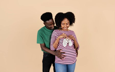 Photo for Happy young black husband hug pregnant wife with big belly in casual, hold small shoes, isolated on beige studio background. Waiting for baby together, love, support, family and relationship - Royalty Free Image