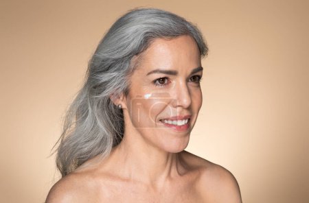 Photo for Portrait of beautiful senior woman with cream on cheek looking aside and smiling, applying hydration moisturizer on face, beige studio background. Beauty day concept - Royalty Free Image