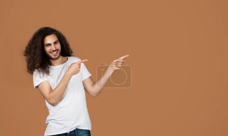 Photo for Special Offer. Portrait Of Happy Middle Eastern Young Man Pointing Fingers Aside At Free Space Standing Over Brown Studio Background. Guy Showing Empty Mockup Place For Advertisement - Royalty Free Image