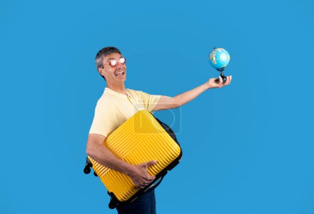 Téléchargez les photos : Globetrotter. Joyeuse mature voyageur homme tenant globe mondial et jaune valise de voyage, posant dans drôle de coeur en forme de lunettes de soleil sur fond bleu studio. Offre vacances d'été - en image libre de droit