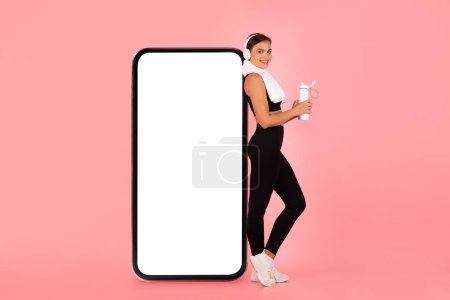 Photo for Fitness Offer. Sporty Young Woman Posing Near Big Blank Smartphone With White Screen, Smiling Athletic Female In Sportswear And Wireless Headphones Recommending New App Or Website, Collage, Mockup - Royalty Free Image