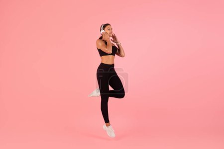 Photo for Portrait Of Young Woman In Sportswear And Wireless Headphones Jumping Over Pink Background, Cheerful Athletic Lady Enjoying Favorite Music While Training, Full Length Shot With Copy Space - Royalty Free Image