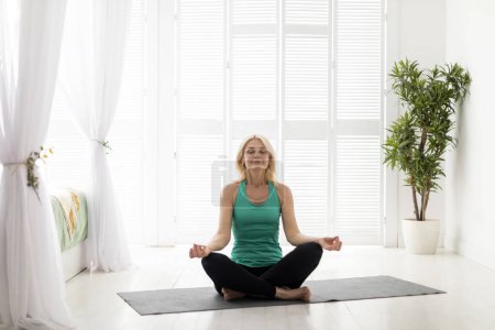 Photo for Morning Meditation. Portrait of calm beautiful senior woman meditating at home, relaxed mature female sitting in lotus position with closed eyes, practicing yoga in light living room, copy space - Royalty Free Image