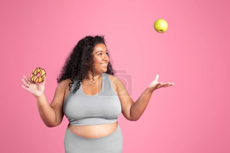 Photo for Excited african american overweight lady in sportswear tossing green apple, chooses between donut or fruit isolated on pink background. Diet, body care, sweets and healthy food - Royalty Free Image