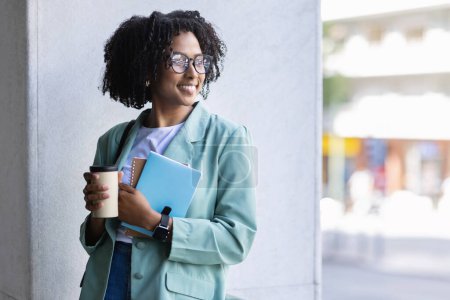 Téléchargez les photos : Joyeux attrayant jeune femme noire dans de belles tenues formelles et des lunettes employé posant à l'extérieur à côté de l'immeuble de bureaux, tenant des blocs-notes et tasse en papier, boire du café, espace de copie - en image libre de droit
