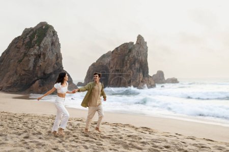 Téléchargez les photos : Voyage de noces. Charmant jeune couple tenant la main et courant le long de la côte de l'océan, s'amusant et appréciant la date sur la plage, espace libre - en image libre de droit