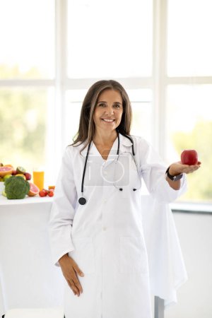 Photo for Cheerful adult european woman doctor nutritionist in white coat offers red organic apple in office clinic interior, vertical. Recommendation health care, proper nutrition, weight loss and diet - Royalty Free Image