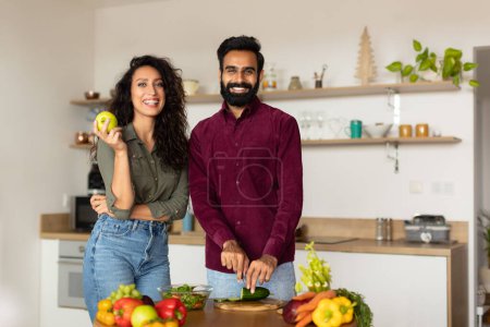 Téléchargez les photos : Portrait de couple heureux du Moyen-Orient posant à l'intérieur de la cuisine, homme cuisinant une salade saine, femme tenant la pomme et souriant ensemble à la caméra, espace libre - en image libre de droit