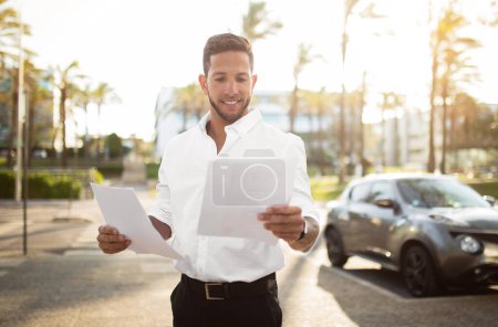 Photo for Positive man employee in formal outfit holding and reading CV, getting ready for job interview, standing outdoors on the street, copy space. Businessman checking documents outside - Royalty Free Image