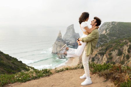 Téléchargez les photos : Vue latérale d'un couple amoureux debout sur une falaise rocheuse et d'un homme tenant une fille dans les airs, profitant d'un moment heureux ensemble et d'une date romantique avec une belle vue sur le littoral, espace libre - en image libre de droit