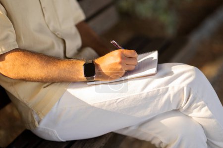Photo for Cropped of man wearing casual outfit sitting on bench on the street or at public park, taking notes in notepad. Unrecognizable guy writing down his thoughts or ideas, planning weekend - Royalty Free Image