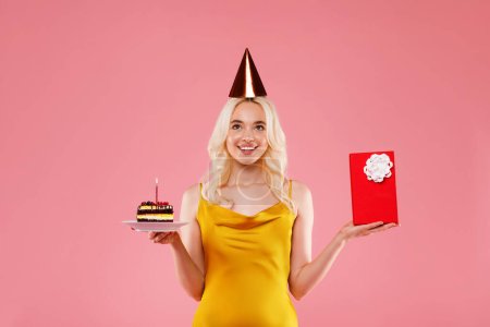 Téléchargez les photos : Joyeux jeune femme portant un chapeau d'anniversaire, tenant gâteau et boîte cadeau rouge, posant isolé sur fond rose, prise de vue en studio. Fête de célébration concept de vacances - en image libre de droit