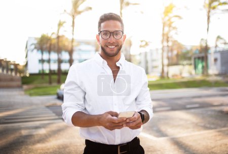 Photo for Happy young european male entrepreneur using cellphone and smiling at camera, checking emails during break outdoors. Cheerful caucasian businessman with phone - Royalty Free Image