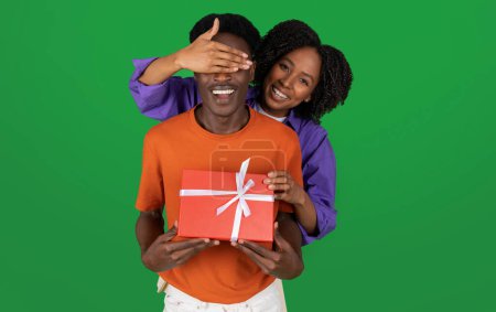 Foto de Sonriente joven rizado mujer afroamericana cierra los ojos al novio, da caja de regalo, aislado en fondo de estudio verde. Sorpresa, amor y relación, cumpleaños, aniversario, celebración de vacaciones - Imagen libre de derechos