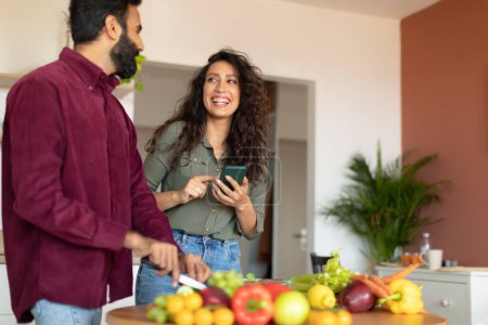 Photo for Happy arab couple cooking together, woman reading recipe on smartphone while man cutting vegetables for salad at kitchen interior, copy space. Excited spouses making lunch - Royalty Free Image
